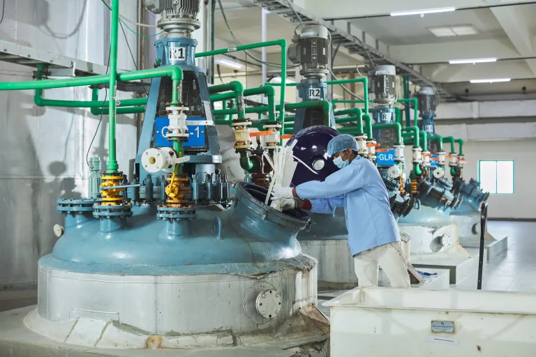 Operator in protective clothing working at a row of reactor vessels on the production floor